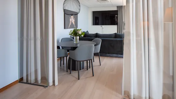 Et spisebord med fire grå stole står ved siden af en grå sofa. En blomsterbuket er på bordet. Rummet er omgivet af lette gardiner.