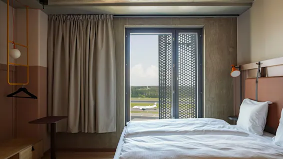 Et moderne hotelværelse med to enkeltsenge og udsigt over en landingsbane gennem et stort vindue. Gardinet dækker delvist vinduet og afslører et fly med "Finnair."