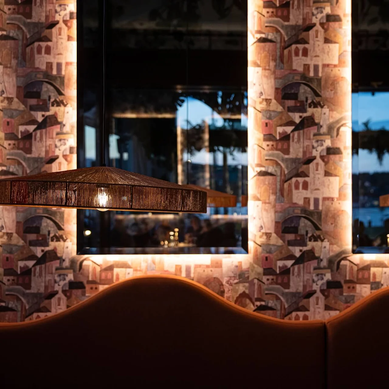 A decorative lamp hangs above a patterned wallpapered wall, softly illuminated. Windows in the background reveal an evening view with glimpses of water and outdoor scenery.