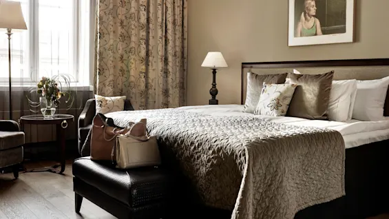 Seng dækket med mønstret quilt, flankeret af puder og sideborde med lamper, i et varmt belyst rum med blomstrede gardiner, kunstværk på væggen og tasker på en bænk.