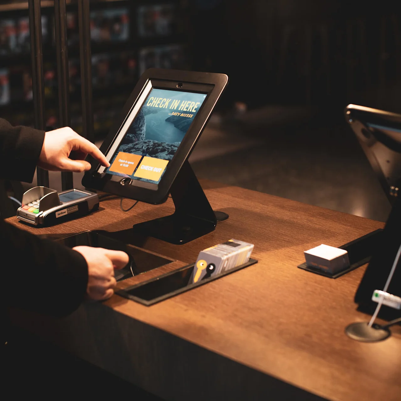 Person interacting with a digital kiosk that reads "CHECK IN HERE - BODY MANOR" and offers options to "Check In" and "Check Out," set in a dimly lit room.