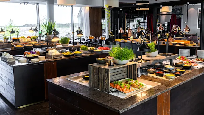 A large buffet table features various dishes including salads, bread, and garnishes. It's surrounded by modern decor with large windows in a well-lit restaurant setting.