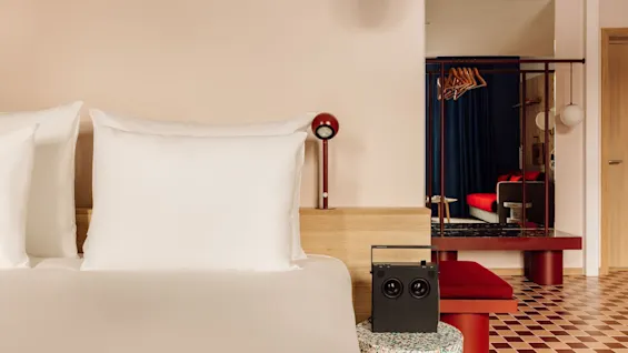 A modern hotel room with a bed in the foreground and a stylish seating area visible through a partition.