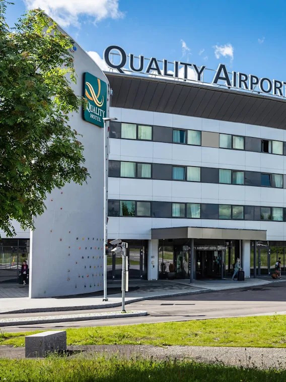 A modern, curved hotel building labeled "Quality Airport Hotel Gardermoen" with a climbing wall, situated amidst trees and flags under a bright blue sky. Text includes "Quality Hotel."