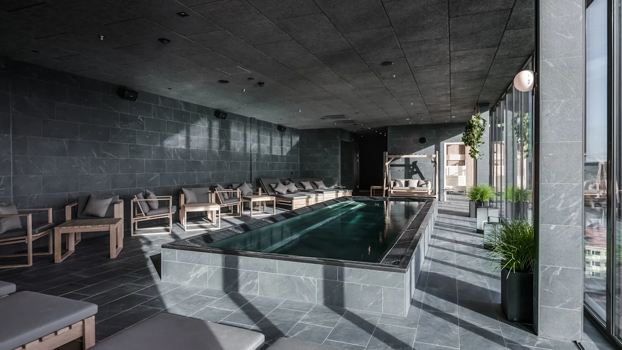 A rectangular indoor swimming pool with clear water, surrounded by cushioned wooden loungers, sits in a modern stone room with large windows and green plants.