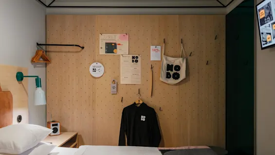 A black hoodie hangs on a wooden wall with pegboard pattern, surrounded by posters and a tote bag. A bed with white sheets and a bedside lamp is nearby.