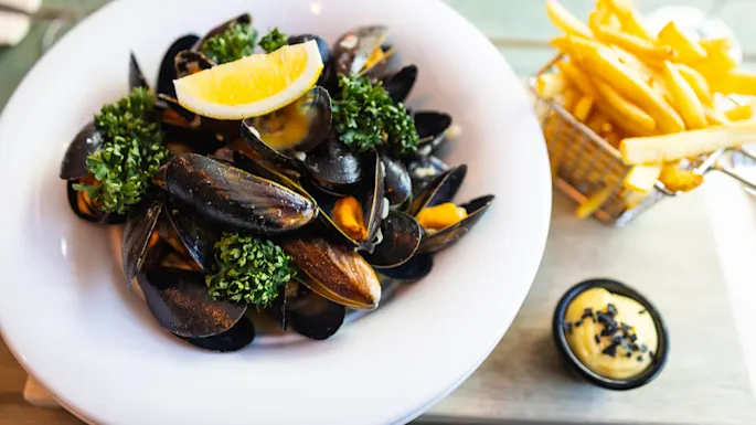 Mussels garnished with lemon and parsley, served in a white bowl, accompanied by a side of French fries in a basket and a small dish of creamy sauce on a wooden table.