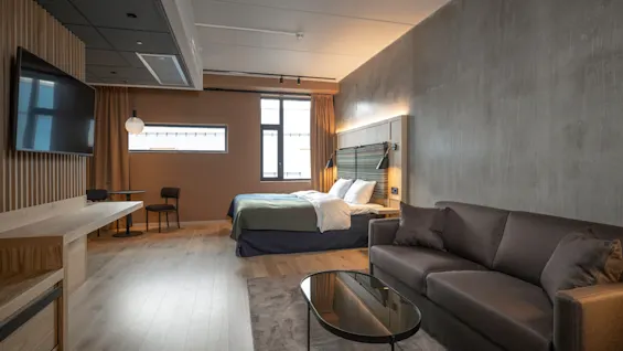 Et pænt indrettet hotelværelse har en redt seng, en sofa og et vægmonteret TV med træpanel. Varm belysning og moderne indretning skaber en hyggelig atmosfære.