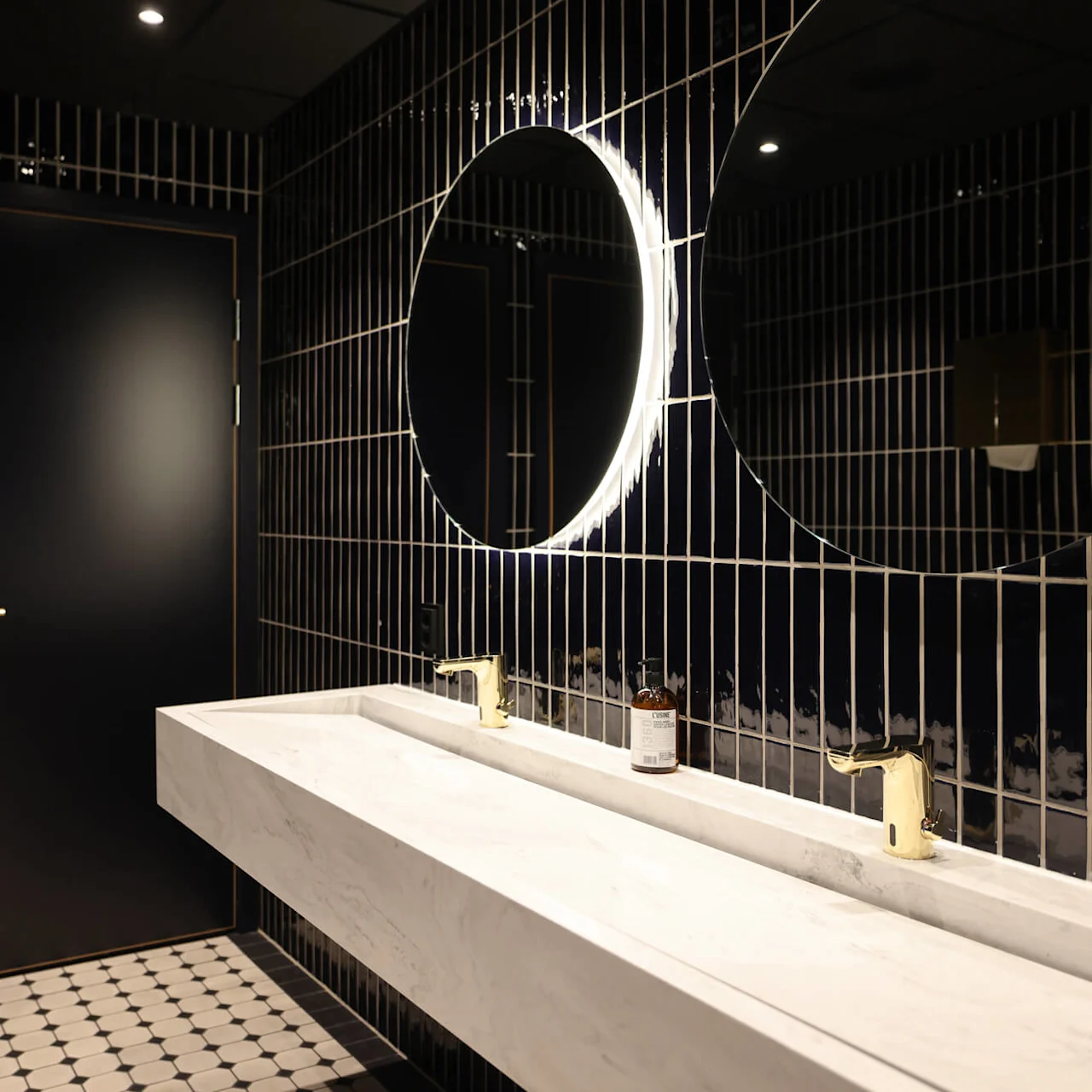 A white marble sink with gold faucets sits below a large illuminated round mirror. Two black doors with gold handles are on a tiled bathroom wall, featuring a geometric floor.