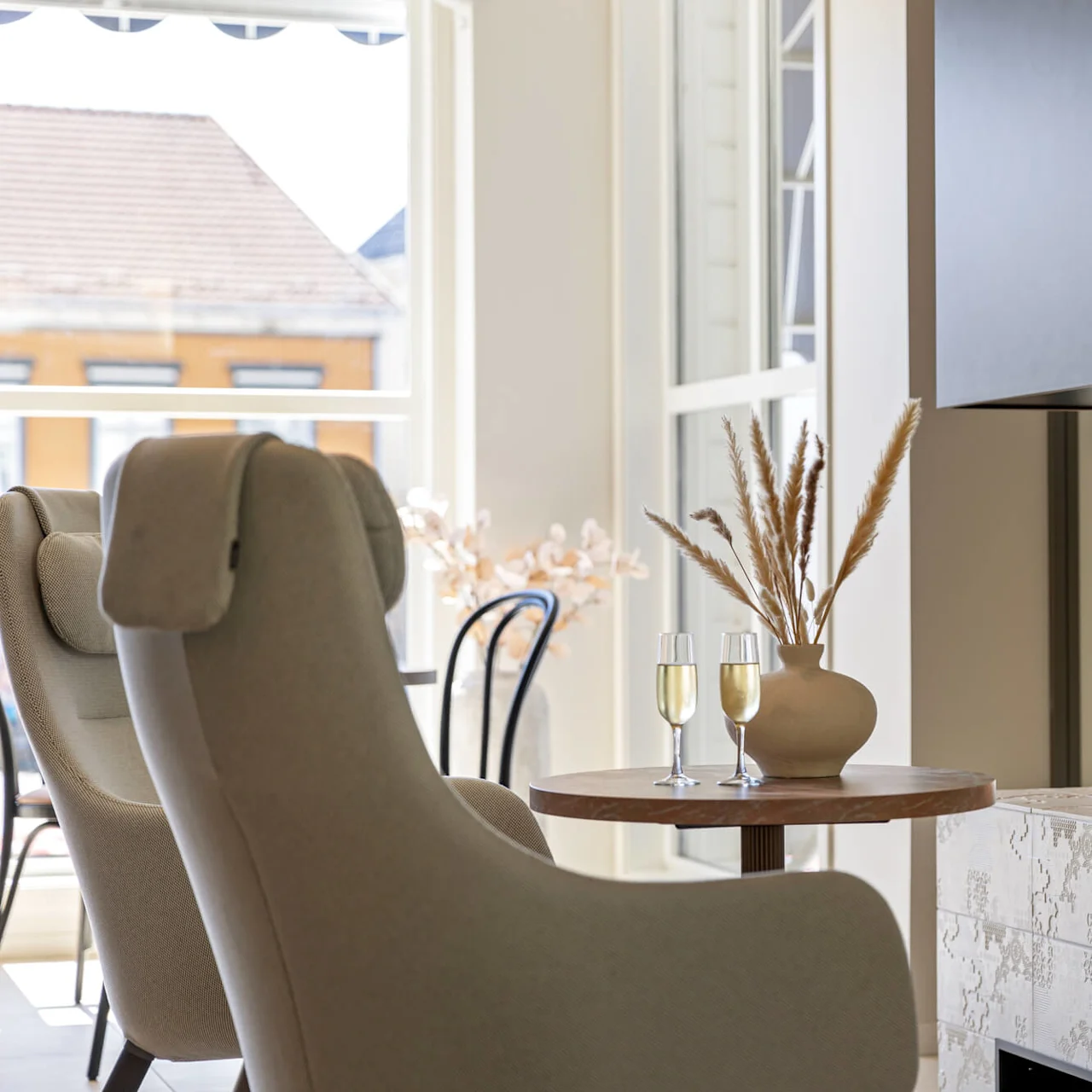 A modern interior with a beige armchair, round table with a vase and glasses, and a fireplace against a window backdrop.