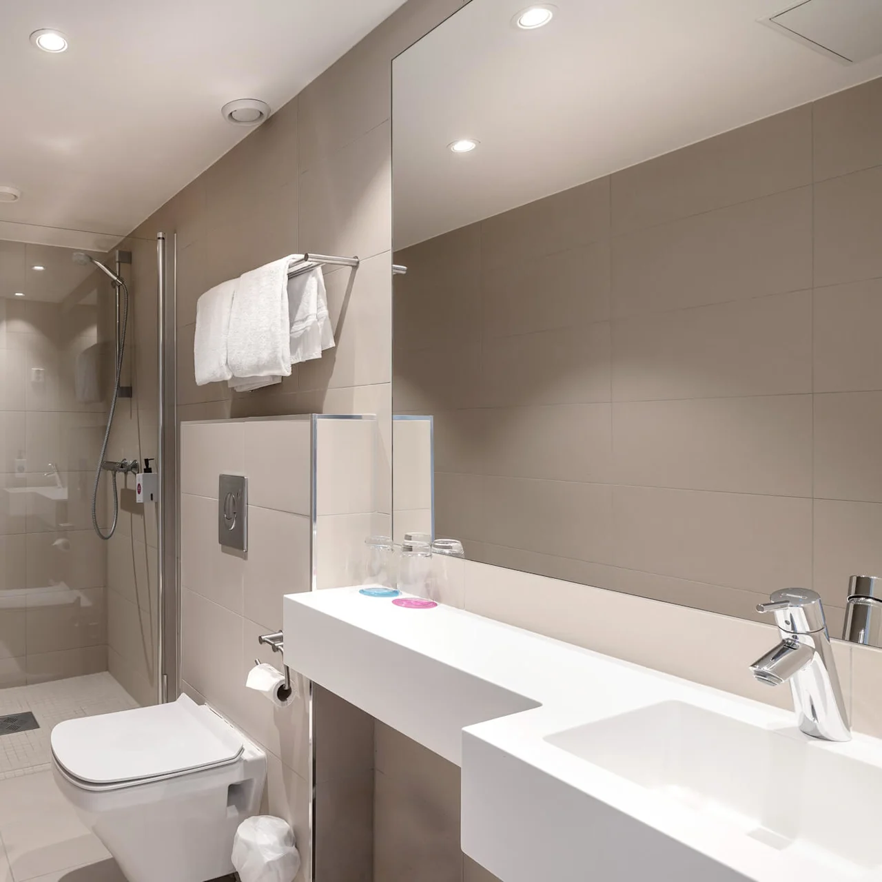 Et moderne bad med en glassinnrammet dusj, vegghengt toalett og strømlinjeformet servant med stor speil. Beige fliser dekker vegger og gulv, og gir et minimalistisk utseende.