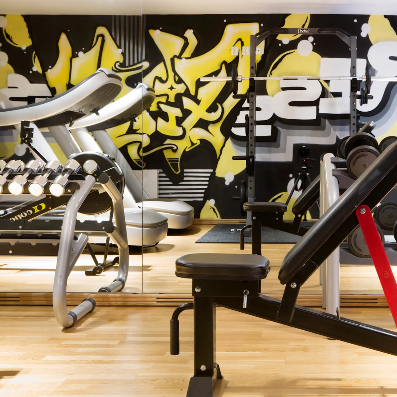 A weight bench faces a mirror across a gym floor, adjacent to a rack filled with dumbbells. Treadmills and vibrant yellow graffiti art decorate the background.