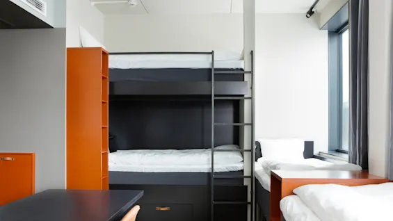 Våningssängar är prydligt arrangerade med vita sängkläder; en stege förbinder sängarna. En enkelsäng står bredvid ett stort fönster, och ett skrivbord med en stol finns i förgrunden.