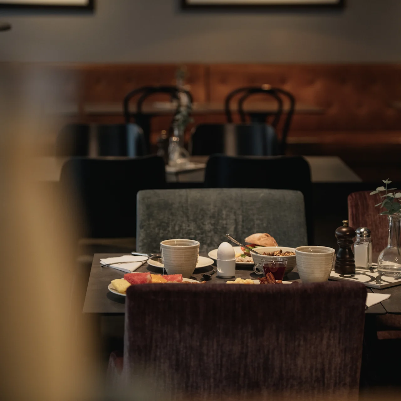 A dining table set with cups, plates, and a teapot, surrounded by upholstered chairs in a cozy restaurant setting.