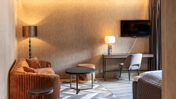 Brun sofa og runde bord i et moderne hotellrom med lamper som lyser opp rommet. En flatskjerm-TV henger på veggen over et skrivebord og en stol. Ingen tekst.