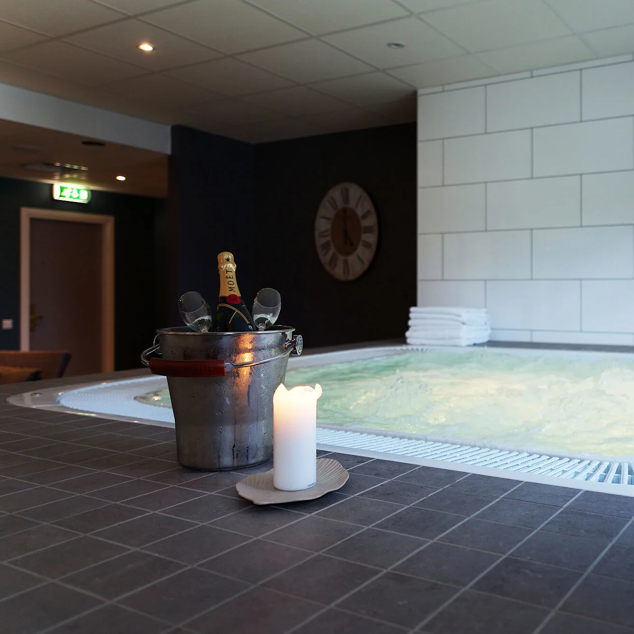 A jacuzzi with bubbling water, a bucket with a champagne bottle, glasses, and a lit candle, implying relaxation in a tiled room with towels stacked nearby.