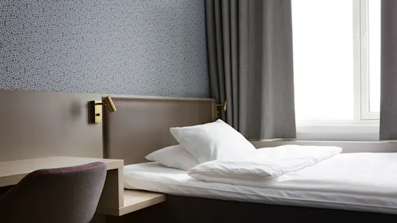A neatly made bed with white bedding in a room with patterned wallpaper and curtains, suggesting a hotel setting.