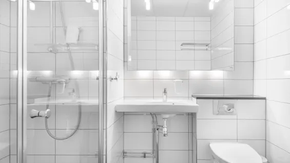 Et lite hvitflislagt baderom med en vask og speil, en dusj med glassdør og en toalett; moderne armaturer og belysning gir et rent, minimalistisk utseende.