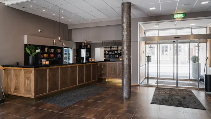 Resepsjonsdisken er laget av tre, med hengelamper over og en vase med en plante. En glassinngangsdør viser gaten utenfor. Utgangsskilt og snacks er synlige.