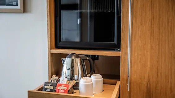 En trähylla innehåller en vattenkokare, tepåsar märkta "Twinings Prince of Wales" och "Twinings English Breakfast", samt två vita koppar. En liten kyl är synlig i närheten.