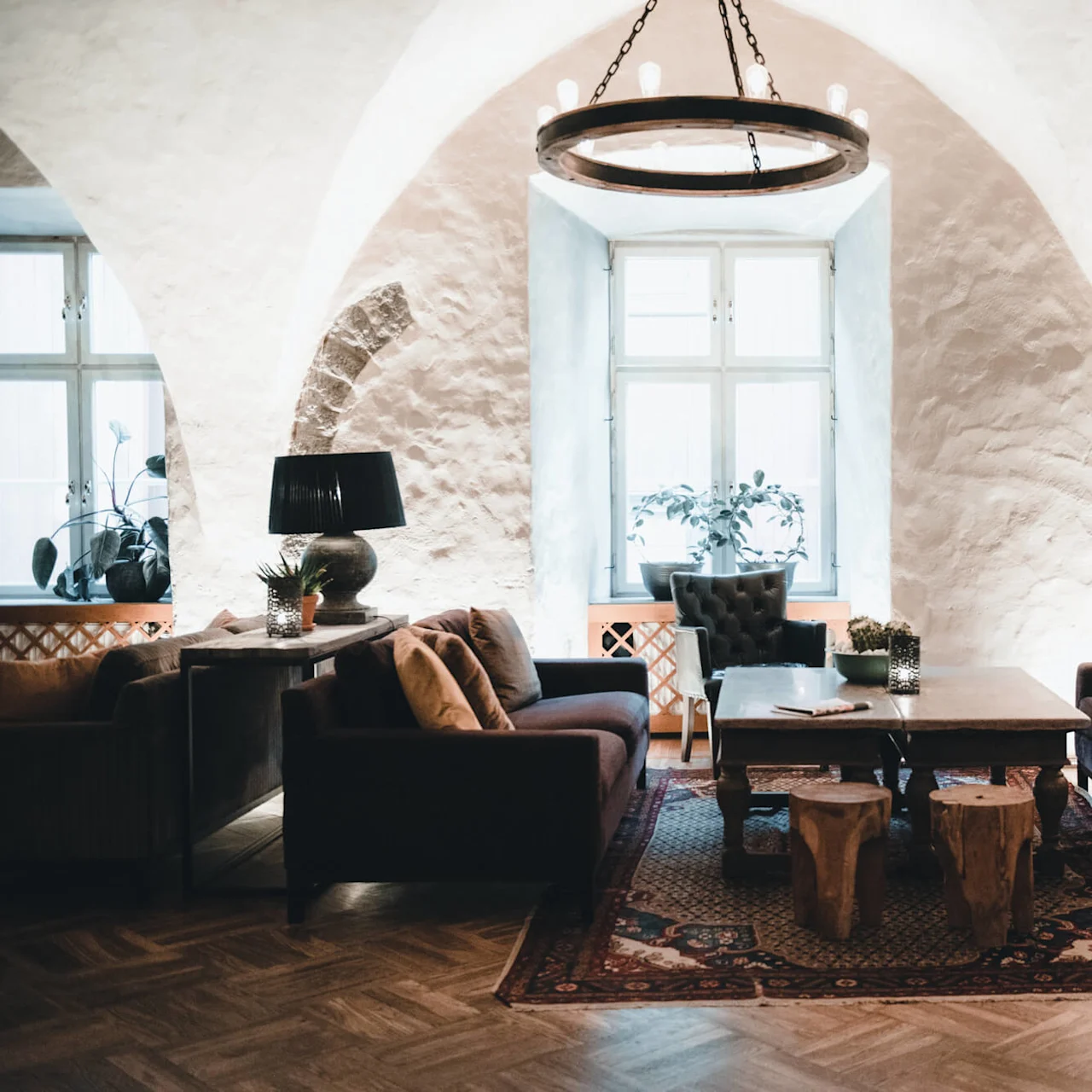 En hyggelig, elegant stue med et træbord omgivet af mørke sofaer, under en rustik lysekrone, med buede stenvægge og store vinduer, der giver naturligt lys.
