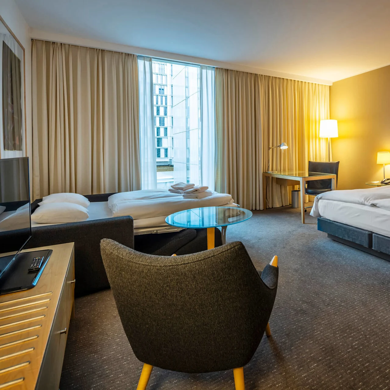 Ett rymligt hotellrum med en stor säng, en bäddsoffa och ett skrivbord. Mjuk belysning och ett fönster med gardiner skapar en mysig atmosfär.