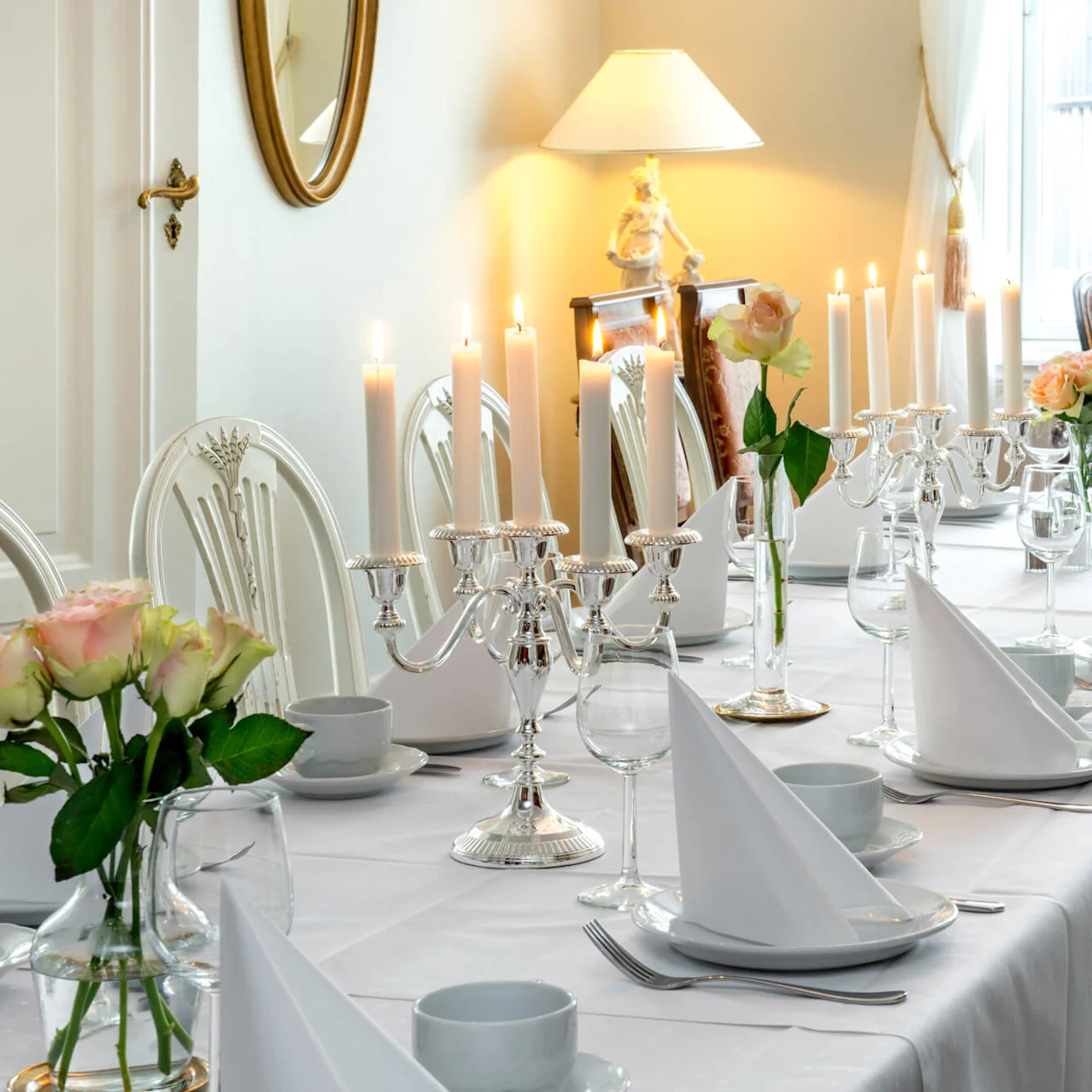 Elegant matservering med tända ljus, fint porslin och färska blommor på en vit duk i ett lyxigt inrett rum.