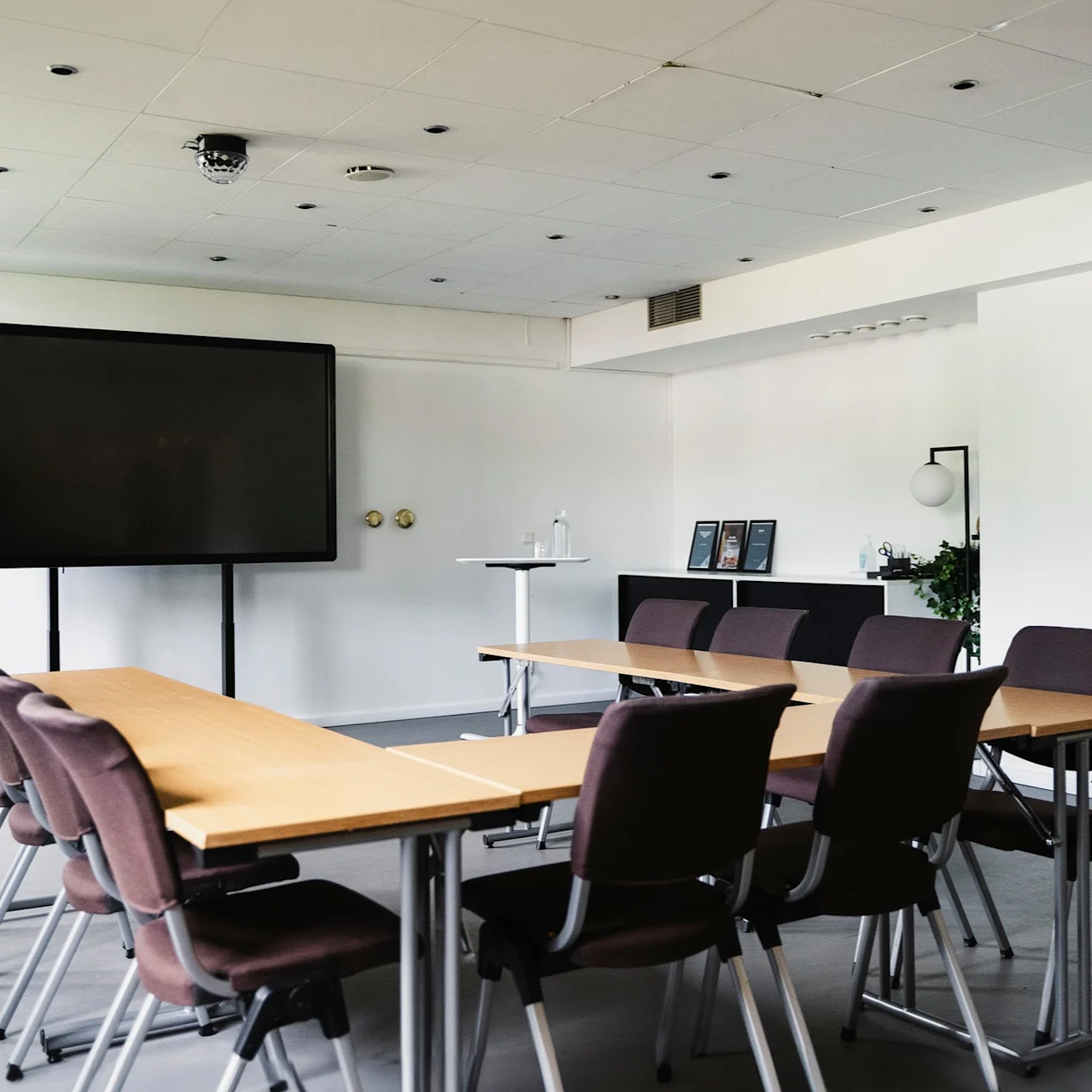 En konferensrum med rader av bruna stolar vända mot en stor svart skärm. Borden är arrangerade i U-form. Vita väggar och modern inredning skapar en minimalistisk miljö.