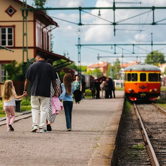 En familj promenerar längs en tågperrong med ett gammaldags tåg på spåren och en stationsbyggnad i bakgrunden.