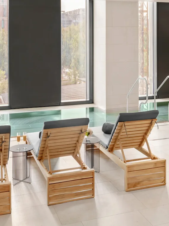 Sunchairs with cushions face an indoor swimming pool, adjacent to large windows. A small table holds glasses of champagne. A robe hangs nearby with text "elements spa."