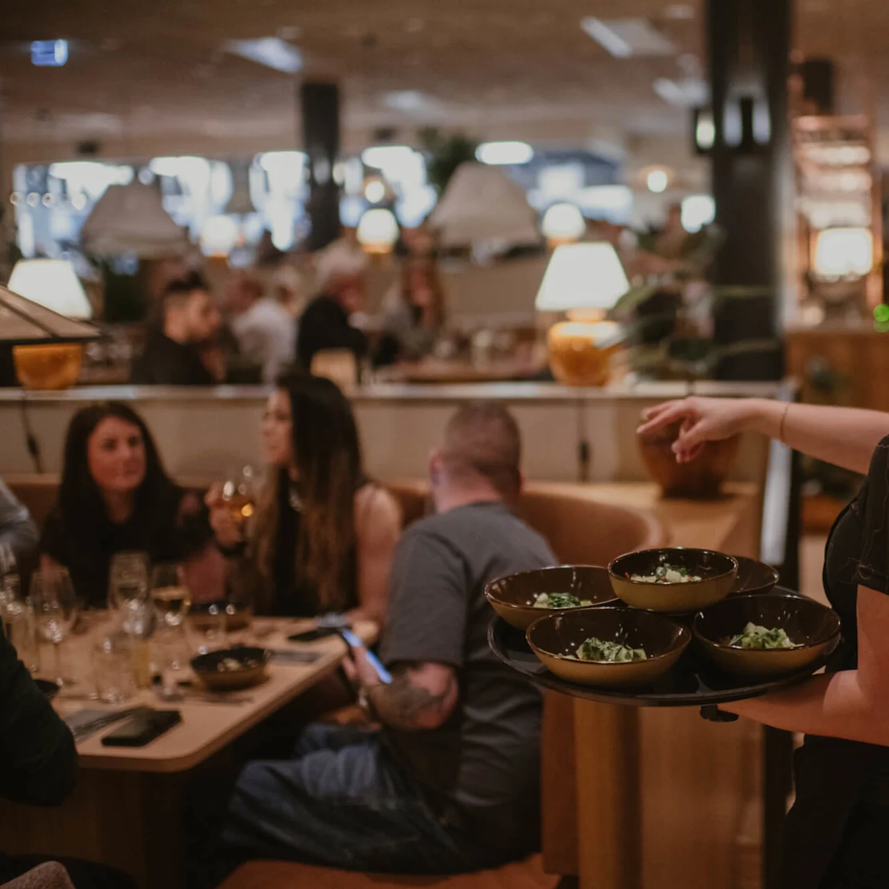 En servitör balanserar skålar på en bricka mot gäster vid bord i en livlig, varmt belyst restaurang.
