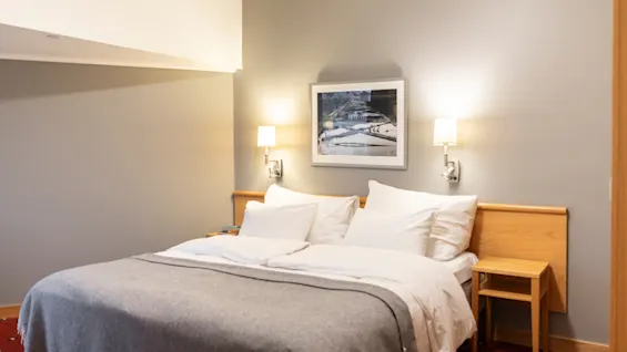 Et koselig hotellrom med en stor seng med hvitt sengetøy og et grått pledd, nattbord og vegglamper.