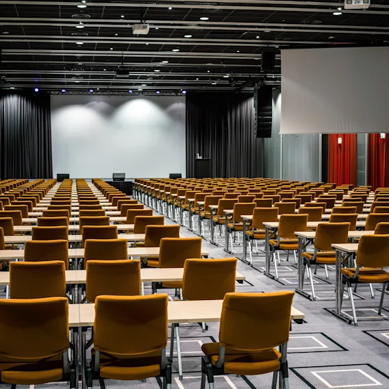 Rader av tomme oransje stoler vender mot en stor, tom skjerm i et romslig konferanserom med mønstret teppe og strukturert belysning.