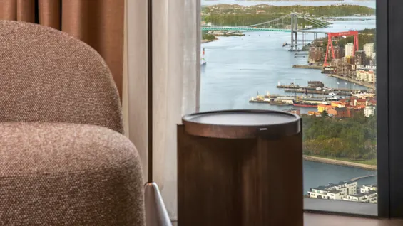 En rund, texturerad fåtölj står nära ett litet träbord, bredvid stora fönster. Utanför syns en flod med en bro och industriellt landskap under en molnig himmel.