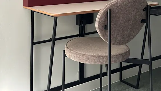 Pult med en blå lampe plassert på en lys treoverflate, sammen med en beige stol i et minimalistisk rom med enkle vegger.