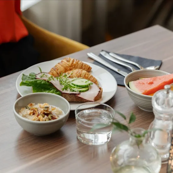Et hotelmorgenbord med et stykke belagt smørrebrød, en croissant, en skål granola, frisk vandmelon og et glas vand.