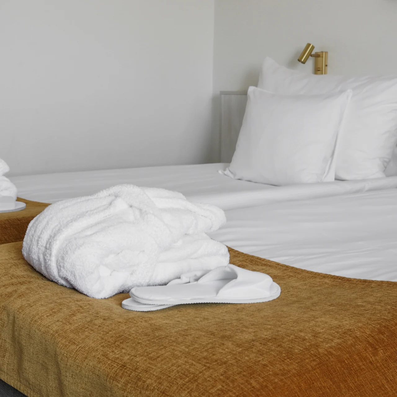 A neatly made bed with white sheets and pillows holds folded bathrobes and slippers on brown blankets. The setting is a minimalistic room with a white wall.