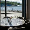 A set dining table with glasses and cutlery, overlooking a scenic waterfront with green hills. Text on window: THE SOCIAL.