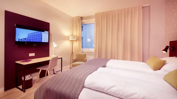Et lyst moderne hotellrom med en pent oppredd seng, et skrivebord, en stol og en flatskjerm-TV på veggen, innredet i varme toner.