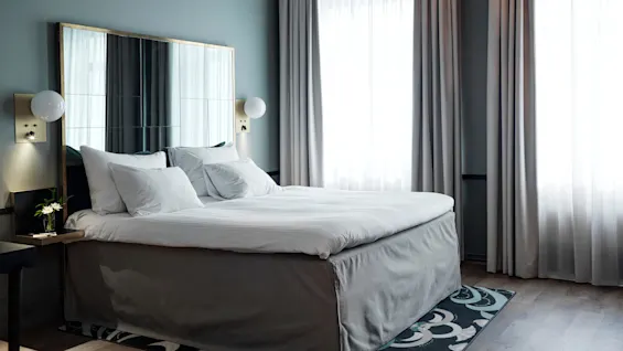 A neatly-made bed with white linens stands in a modern room. Soft daylight filters through sheer curtains. A vase with flowers is on the bedside table beside a decorative lamp.