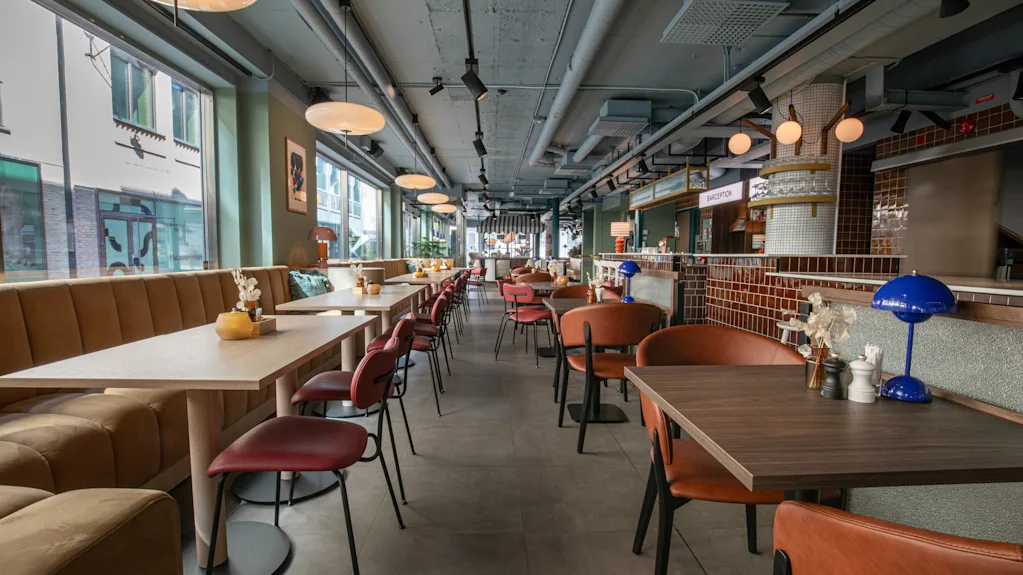 Et moderne og indbydende restaurantinteriør med borde, stole og båse langs store vinduer. En receptionsdisk er synlig.