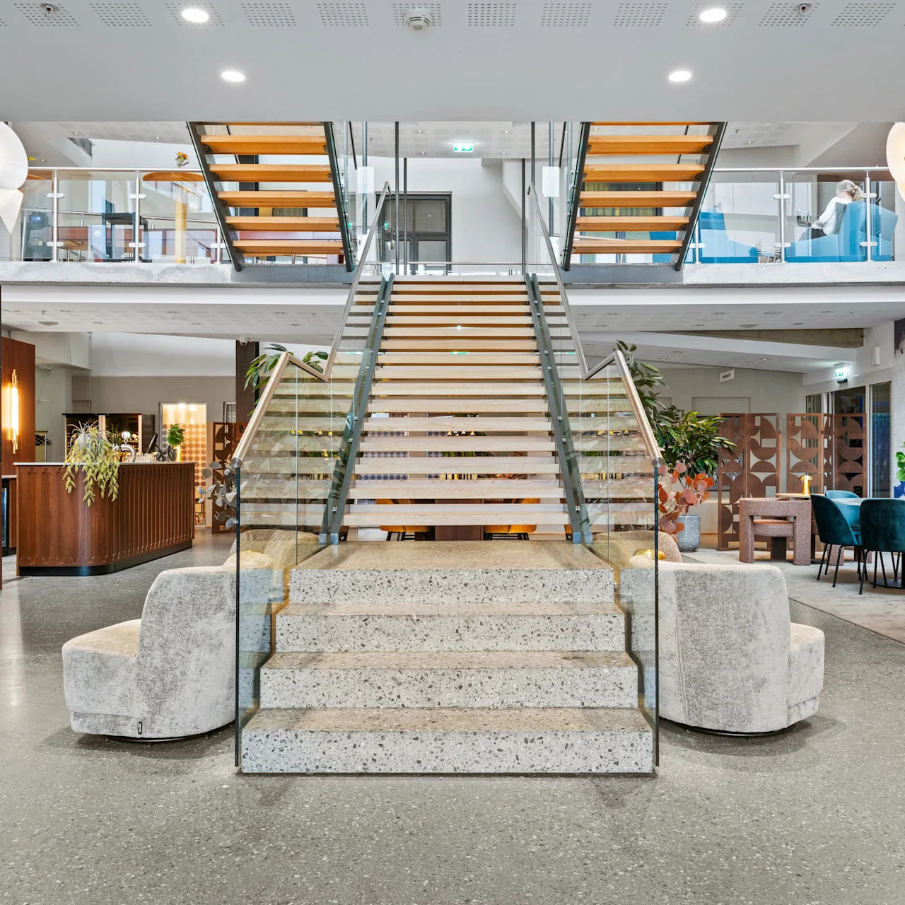 Trappa reser sig i modern lobby med glasräcken, omgiven av sittplatser och växter. Olika belysningsarmaturer lyser upp och skapar en välkomnande atmosfär.