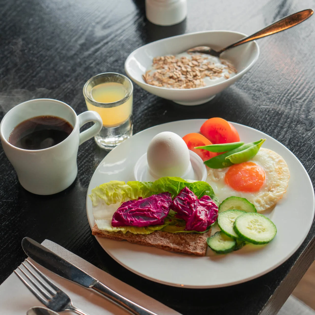 Breakfast plate with boiled egg, fried egg, vegetables, and rye crispbread beside coffee, juice, yogurt, and cereal. Set on a dark wooden table with utensils and napkin.