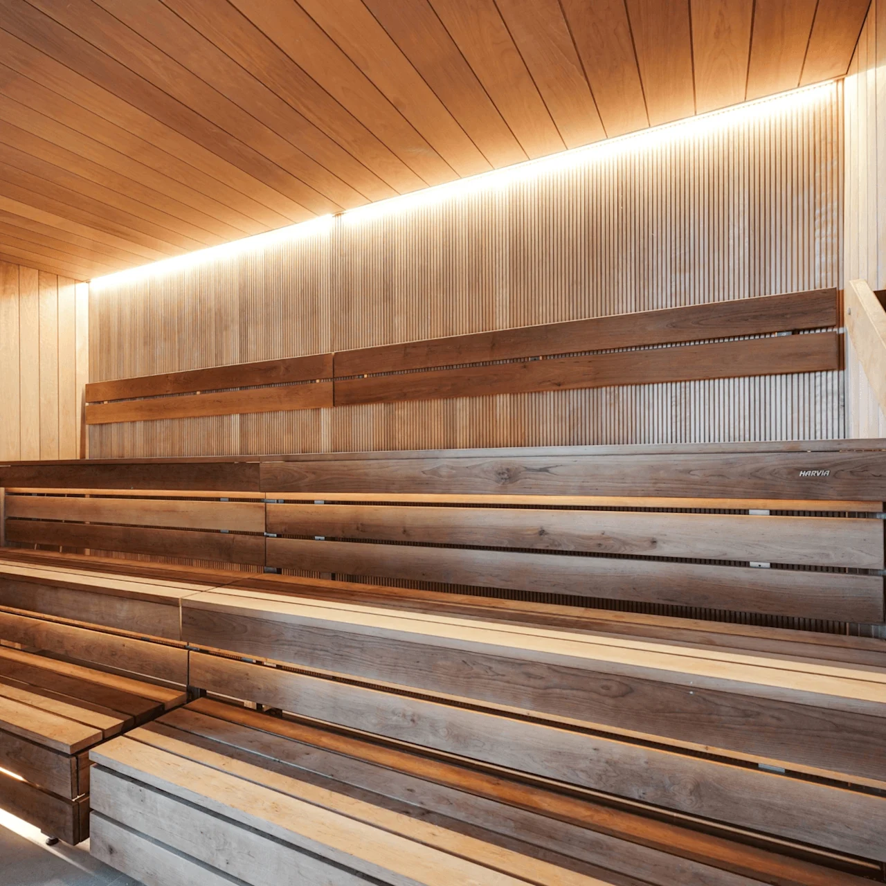 A wooden sauna features tiered benches, illuminated by soft lighting, in a serene setting. A Harvia heater filled with stones sits in the corner.