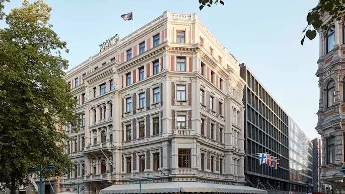 Elegant hjørnebygning med en kafé på gateplan, i en urban omgivelse med trær og klar himmel.