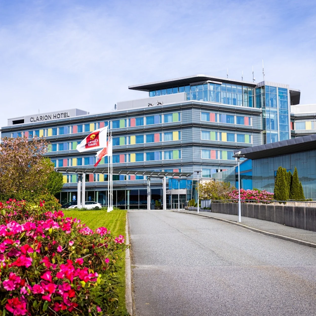 A modern, multi-story hotel labeled "CLARION HOTEL" stands amidst landscaped gardens with vibrant pink flowers. The scene is bright, under a clear blue sky. Nearby, a flag displays a hotel logo.