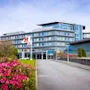 Et moderne fleretasjes hotell med påskriften "CLARION HOTEL" står blant velstelte hager med livlige rosa blomster. Scenen er lys under en klar blå himmel. Et flagg i nærheten viser hotellets logo.