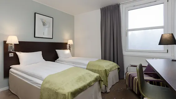 Two neatly made twin beds with green throws are beneath wall lamps in a softly lit hotel room with a window, curtains, and a desk with a chair.