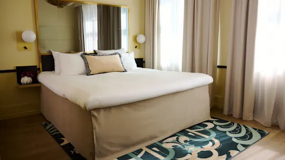 A large bed with white and beige pillows is neatly made, positioned against a wall with a mirror. The room has light-colored curtains and a patterned carpet on wood flooring.