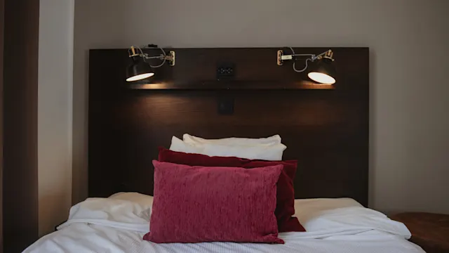 Säng med röda och vita kuddar står mot en mörk träpanel med två justerbara lampor. I en neutralt färgad miljö skapas en mysig och inbjudande atmosfär.